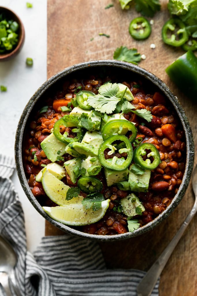 Overhead view of a grey bowl filled with slow cooker vegan lentil chili topped with jalapeño, avocado, and limes.