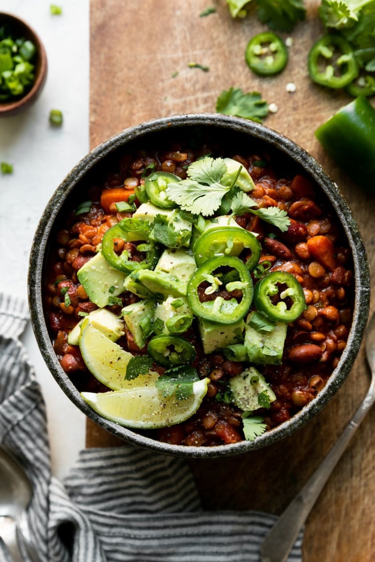 A bowl of slow cooker vegan chili topped with lime wedges and sliced jalapeno. 