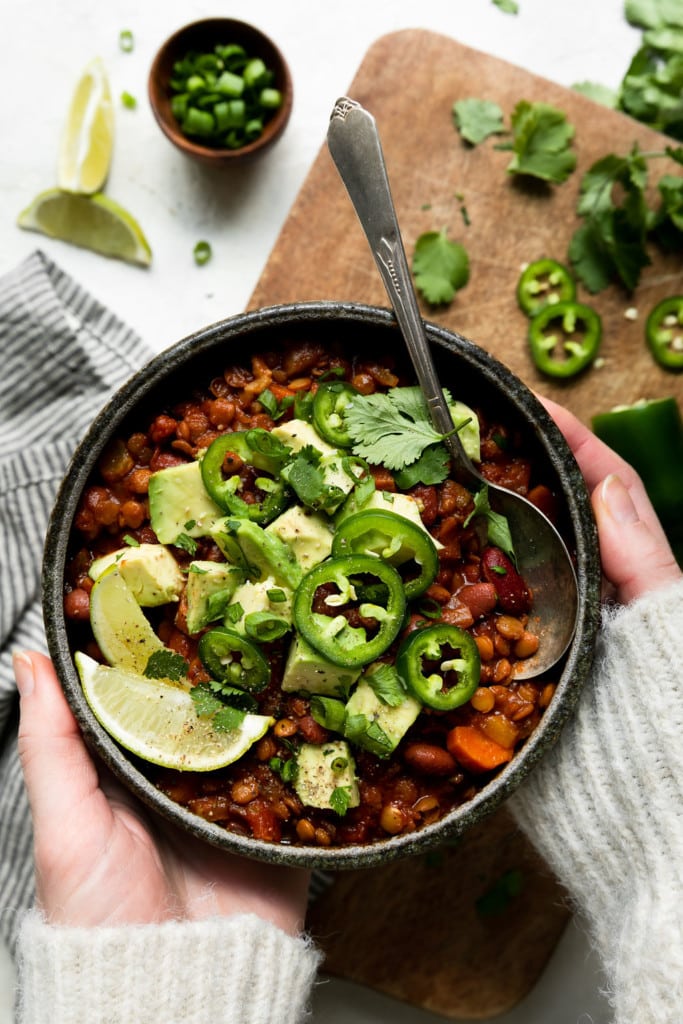 Overhead view of vegan lentil chili in a grey bowl topped with jalapeno slices, avocado, and lime wedges with bowl being held in two hands