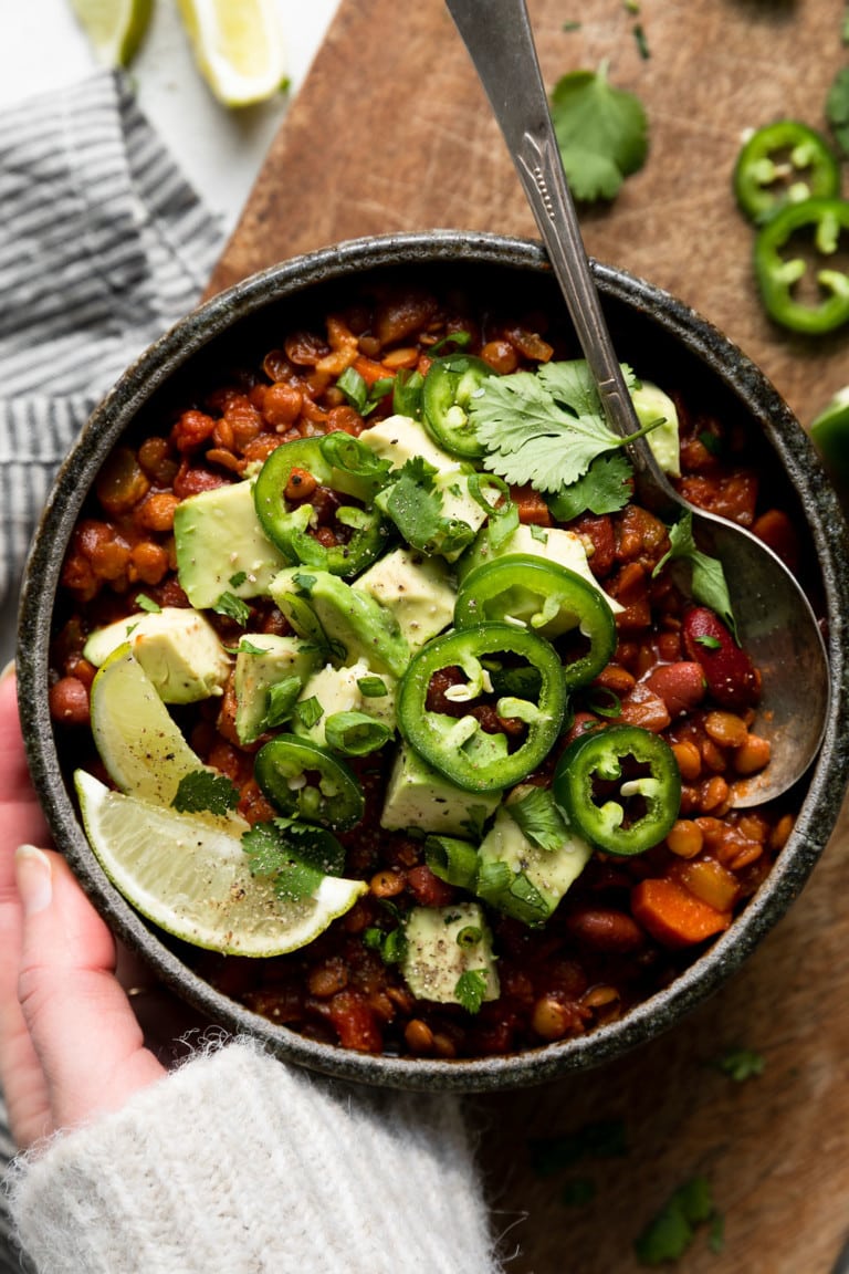 Overhead view of hearty slow cooker vegan chili with lentils in a black bowl topped with avocados, jalapenos, cilantro, and lime wedges. 