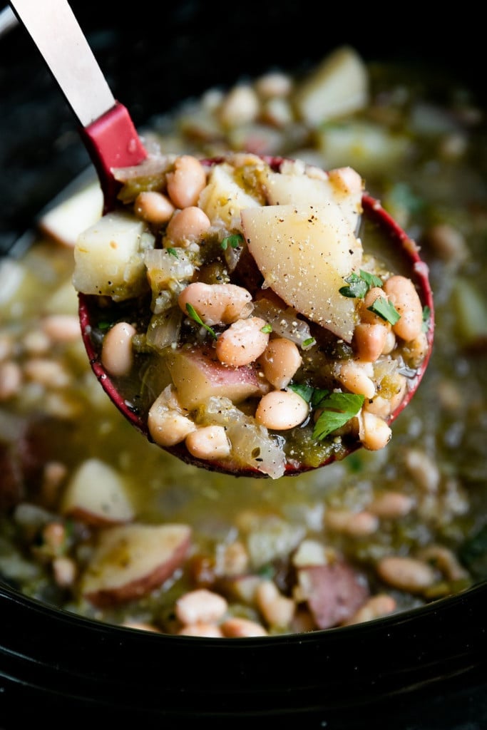 A ladle lifting up a serving a vegan green chili stew over a slow cooker