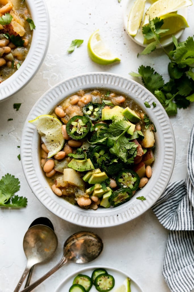 Overhead view of white bowl filled with slow cooker green chili stew topped with avocado, sliced jalapeño, and lime wedges.