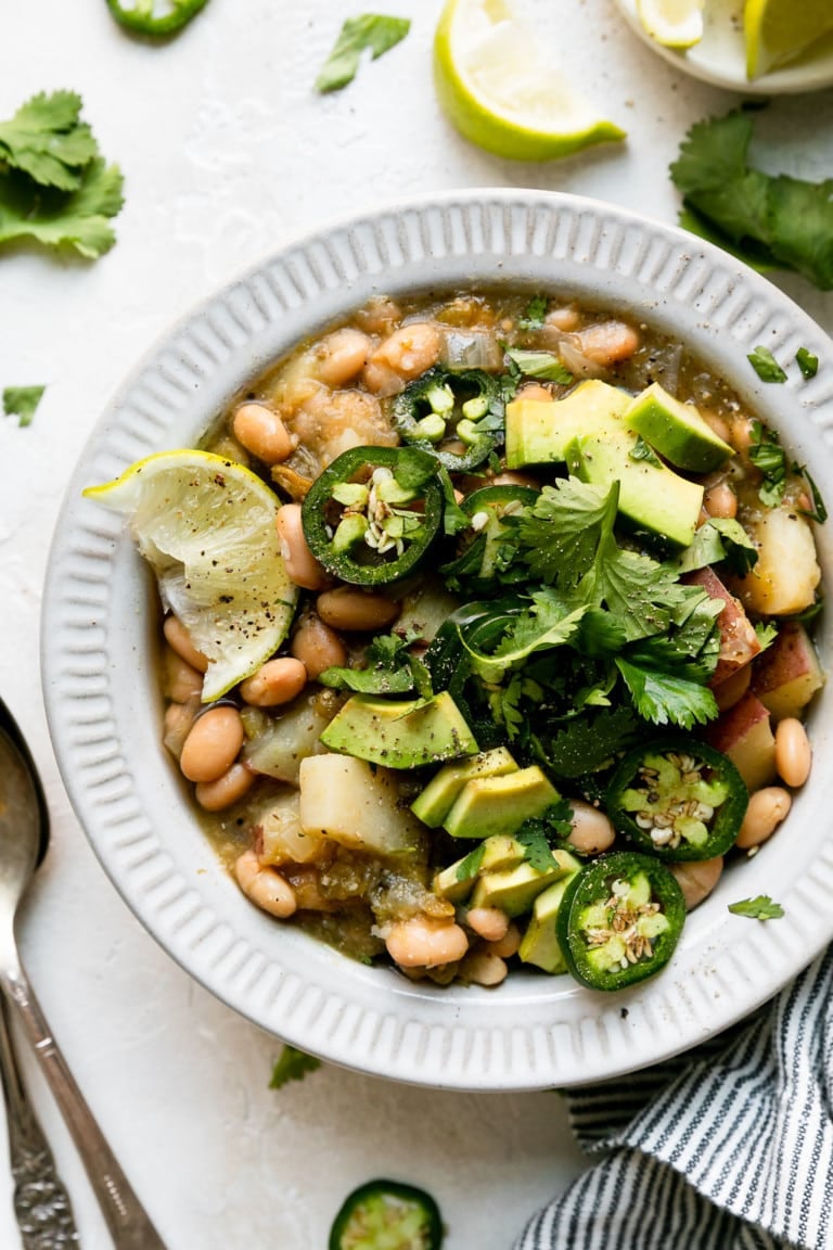 Close up overhead view of slow cooker green chile stew with white beans and potatoes in a white bowl and topped with jalapenos, avocados, and lime wedges. 