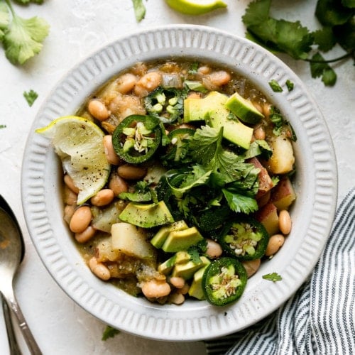 Overhead view of slow cooker green chile stew in a white bowl topped with fresh cilantro, avocado, and jalapeño.