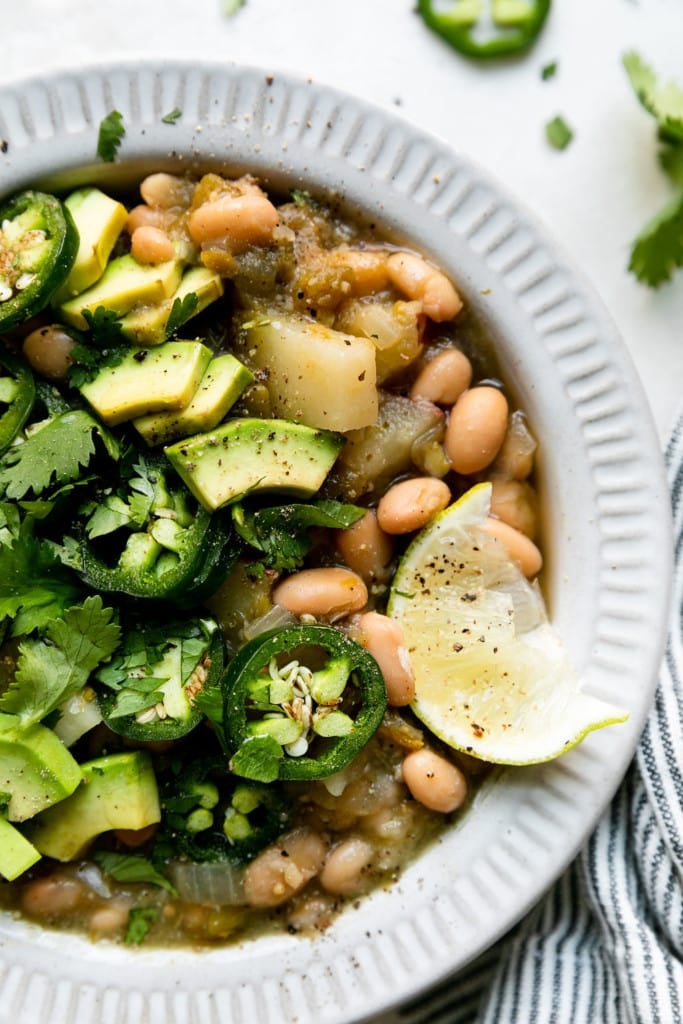 Close up view of slow cooker green chili stew in a white bowl with white beans and potatoes and topped with cilantro, avocado, and sliced jalapeño.