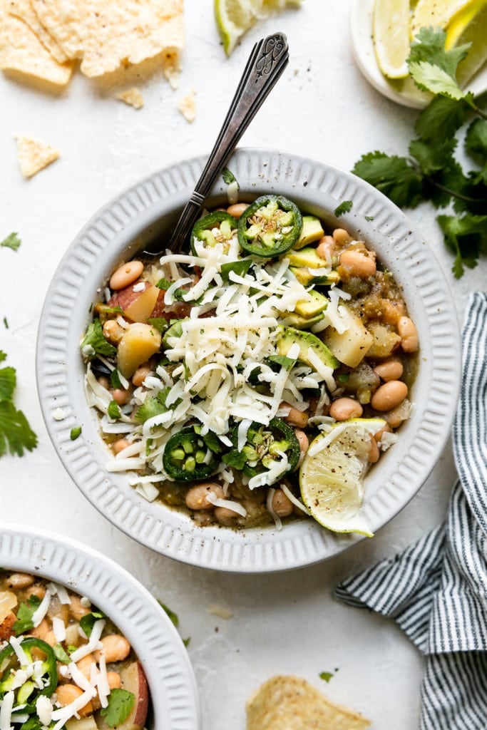 A white bowl filled with slow cooker green chile verde stew with white beans and potatoes topped with jalapeño, avocado, and white shredded cheese.