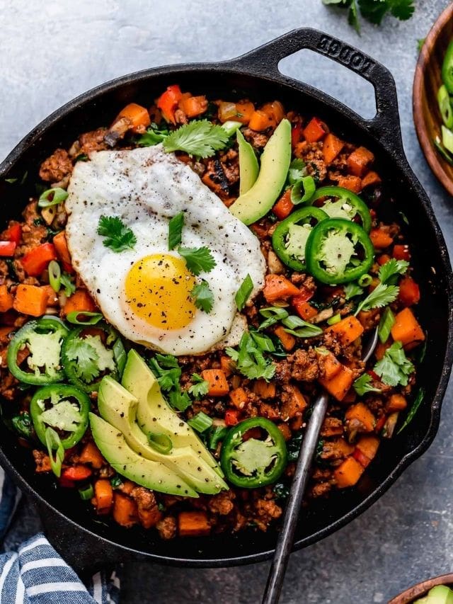 Overhead view cast iron skillet filled with Tex-Mex Sweet Potato Hash, topped with fried egg, avocado slices, and jalepeño slices.
