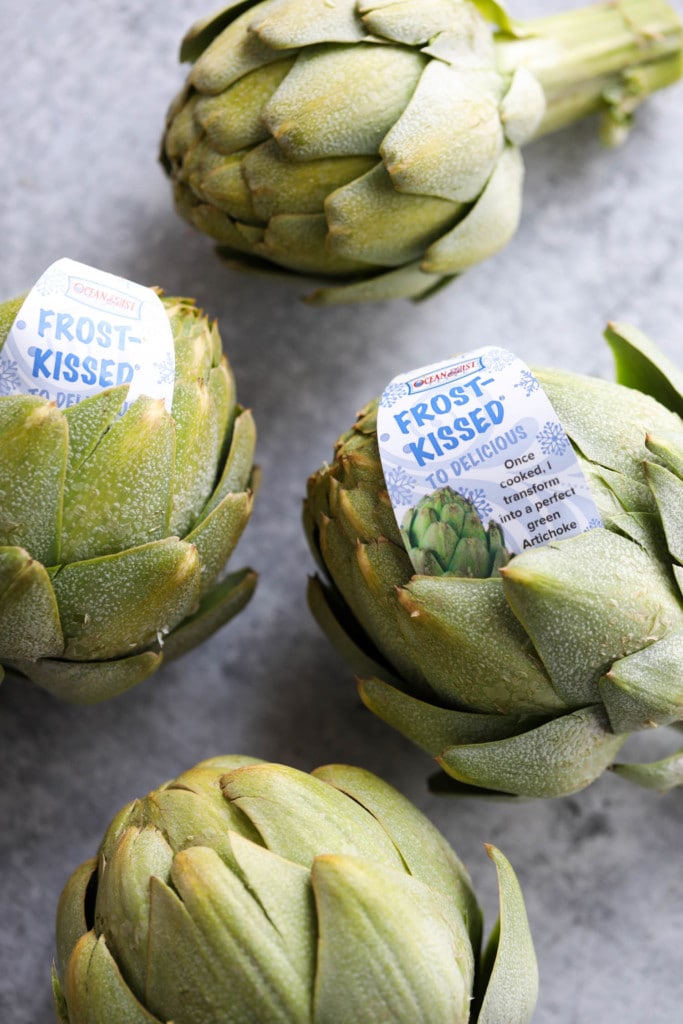 Three whole frost-kissed artichokes arranged together on a cement-counter.
