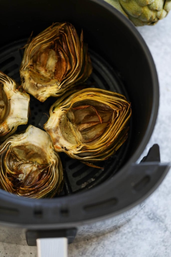 Four artichoke halves in an air fryer basket cut side up, golden brown from air frying.