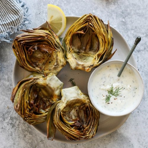 Air Fryer Artichokes on a plate with a small bowl of feta yogurt dip for dipping.