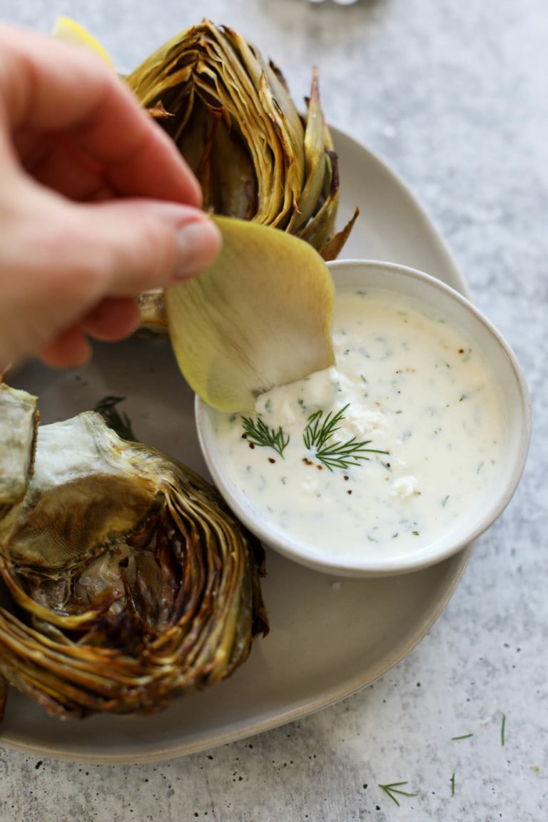A cooked artichoke leaf being dipped into feta yogurt dip from small bowl
