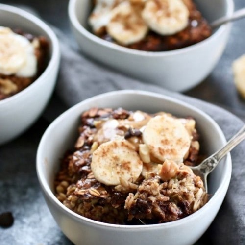 Pieces of banana chocolate chip baked oatmeal in white bowls topped with banana slices and peanut butter.