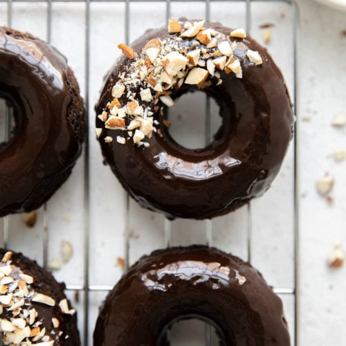 Overhead view of chocolate donuts topped with shiny chocolate icing and crushed almonds, on a wire cooling rack.