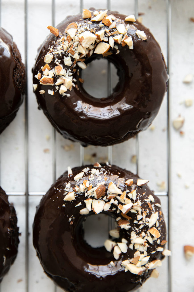 Close up overhead view of baked flourless chocolate donut with shining chocolate icing and crushed almonds on one side of the donut, on a wire cooling rack.