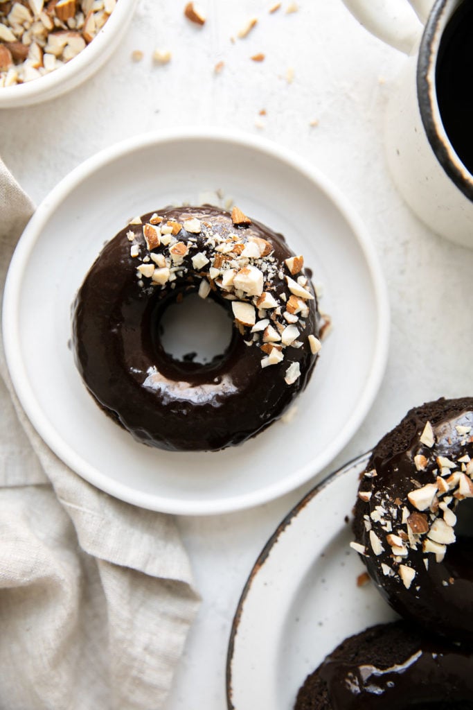 Overhead view of flourless chocolate donut with chocolate icing and crushed almonds sprinkled over half the donut on a small white plate. 