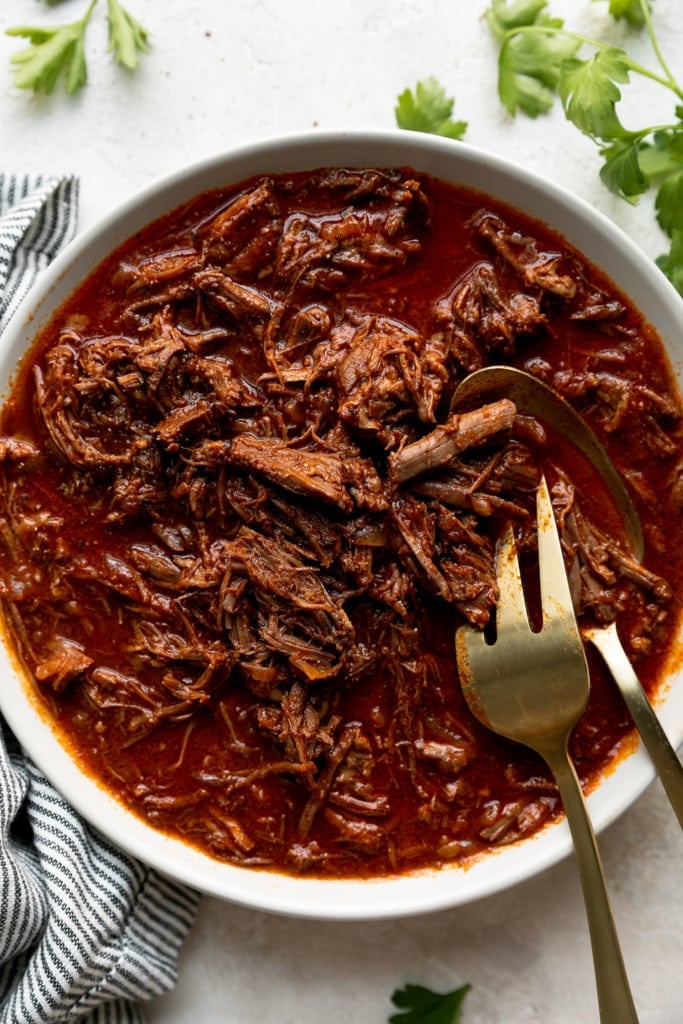 Saucy and juicy shredded beef in bbq sauce served in a white serving bowl with a gold fork and spoon shredding the meat.