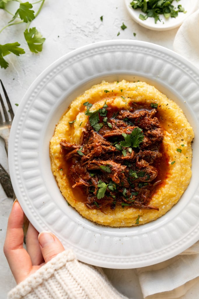 Overhead view of juicy, saucy Instant Pot Shredded Beef served over a bowl of creamy polenta with fresh herbs garnishing top.
