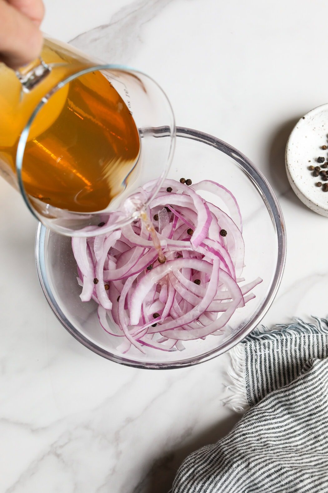 Overhead view of a hand pouring a vinegar mixture over sliced red onions in a glass bowl. 