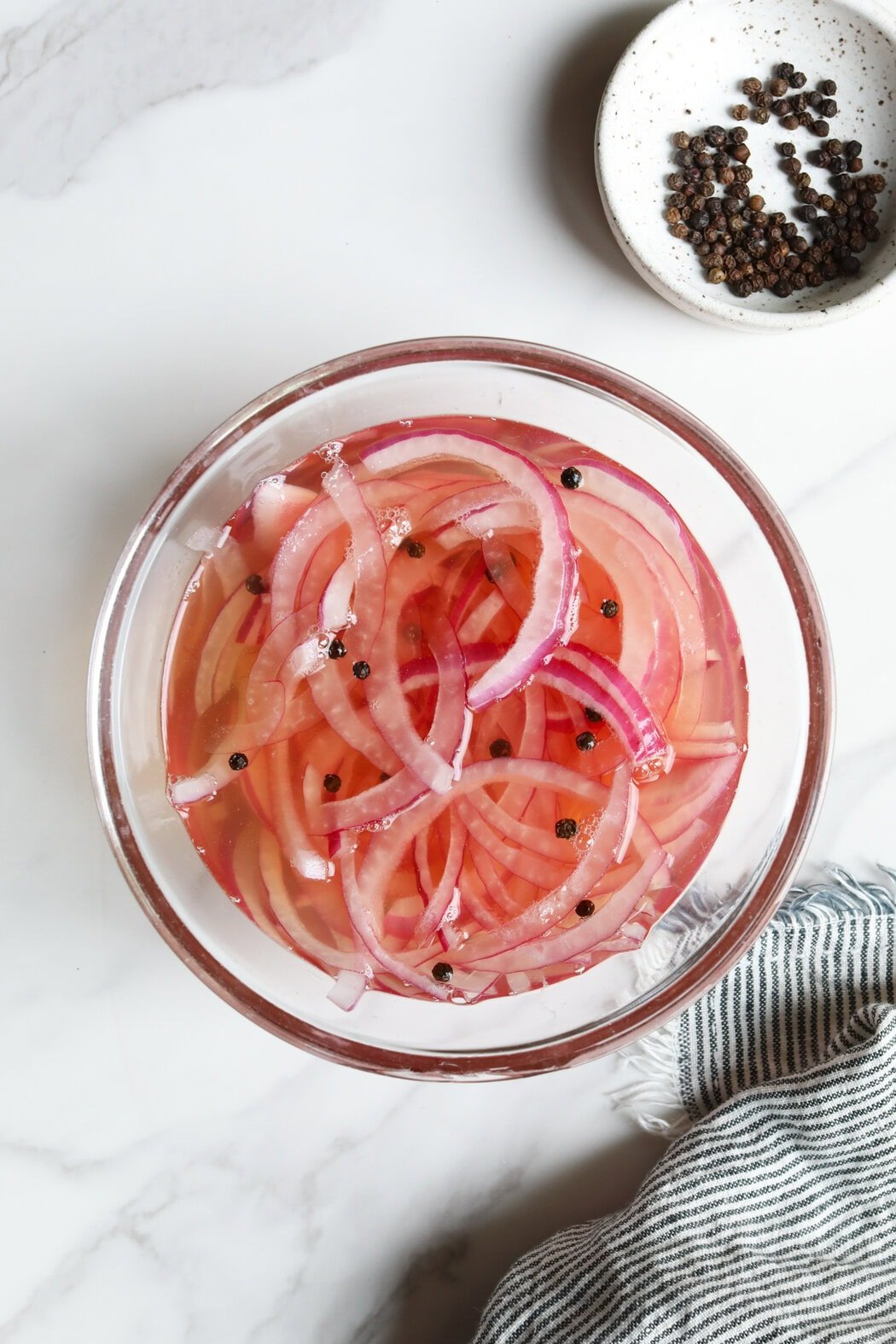 Overhead view of a glass bowl filled with thinly sliced red onions in a vinegar mixture topped with black peppercorns. 