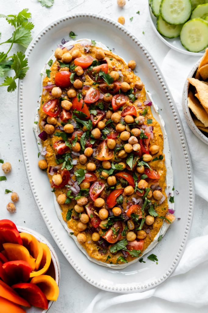 A layered Shawarma dip of Greek yogurt, hummus, Shawarma spices, and topped with chickpeas, tomatoes, and herbs on a white platter.