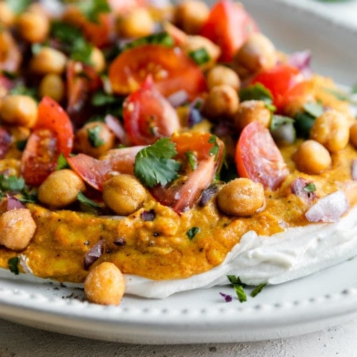 Close up view of layered Shawarma Dip of Greek yogurt, Shawarma seasoned hummus, and chickpeas, tomatoes, and cilantro on a white platter.