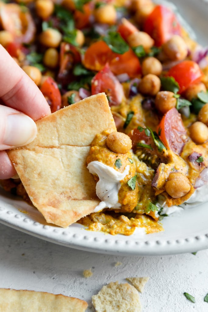 A square of Naan bread scooping up a serving of layered Shawarma dip with roasted chickpeas, tomatoes, and cilantro.