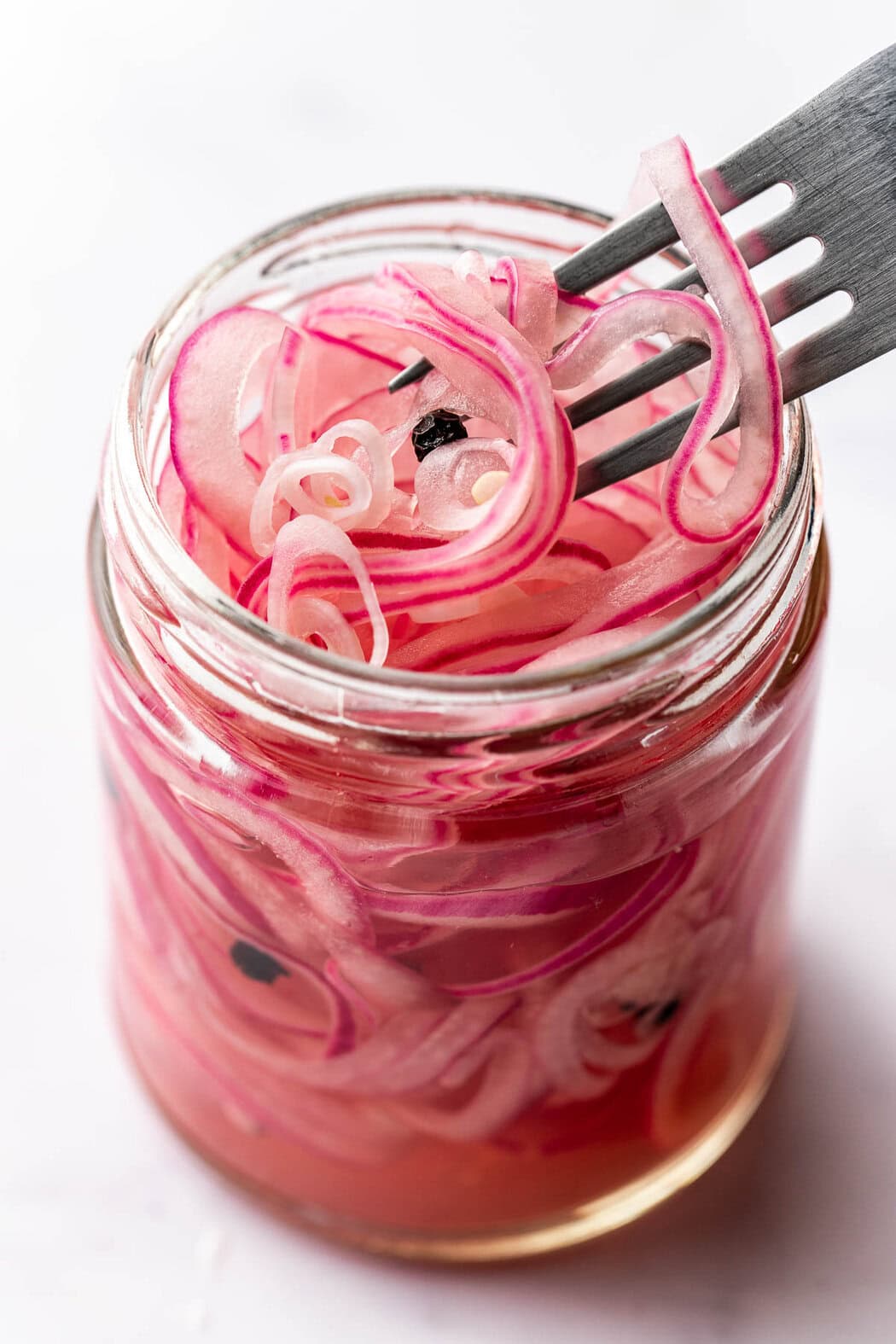 Close up view of a glass jar filled with bright purple Quick Pickled Onions and a fork bringing out some slices. 