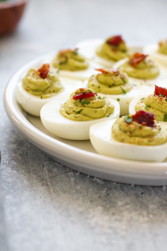 Close up view of creamy filled avocado deviled eggs topped with a small piece of bacon for green eggs and ham recipe.