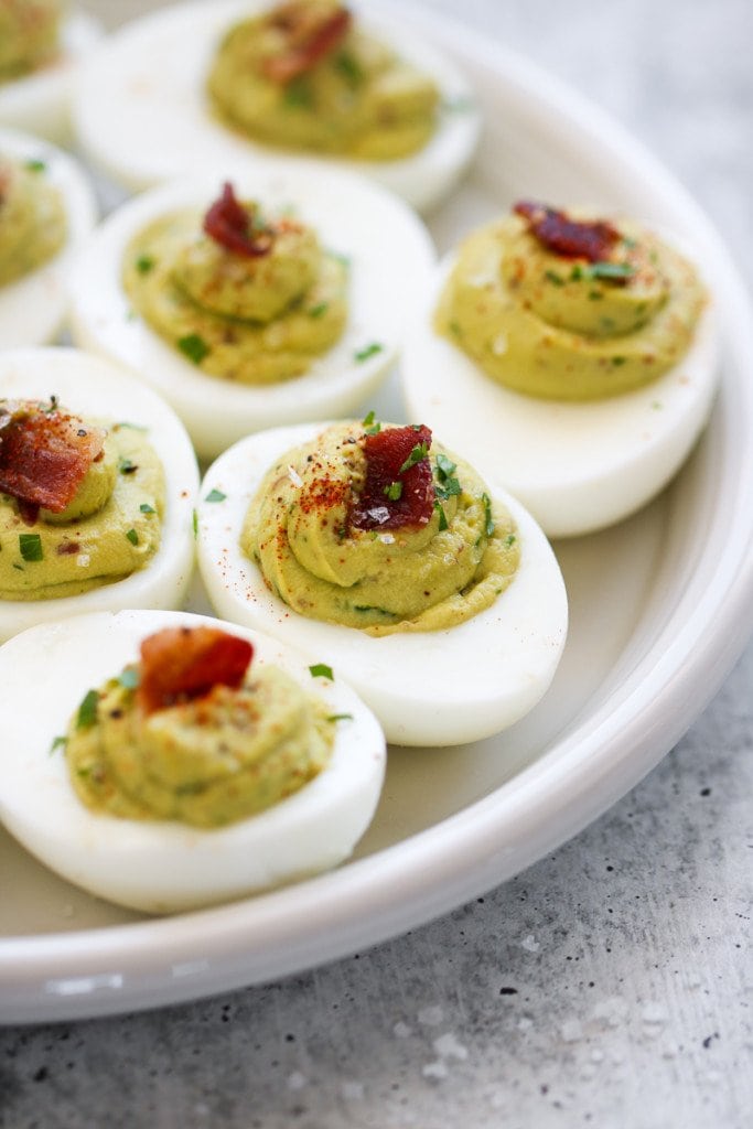 Close up view of avocado deviled eggs topped with bacon and sea salt flakes on a white serving platter.