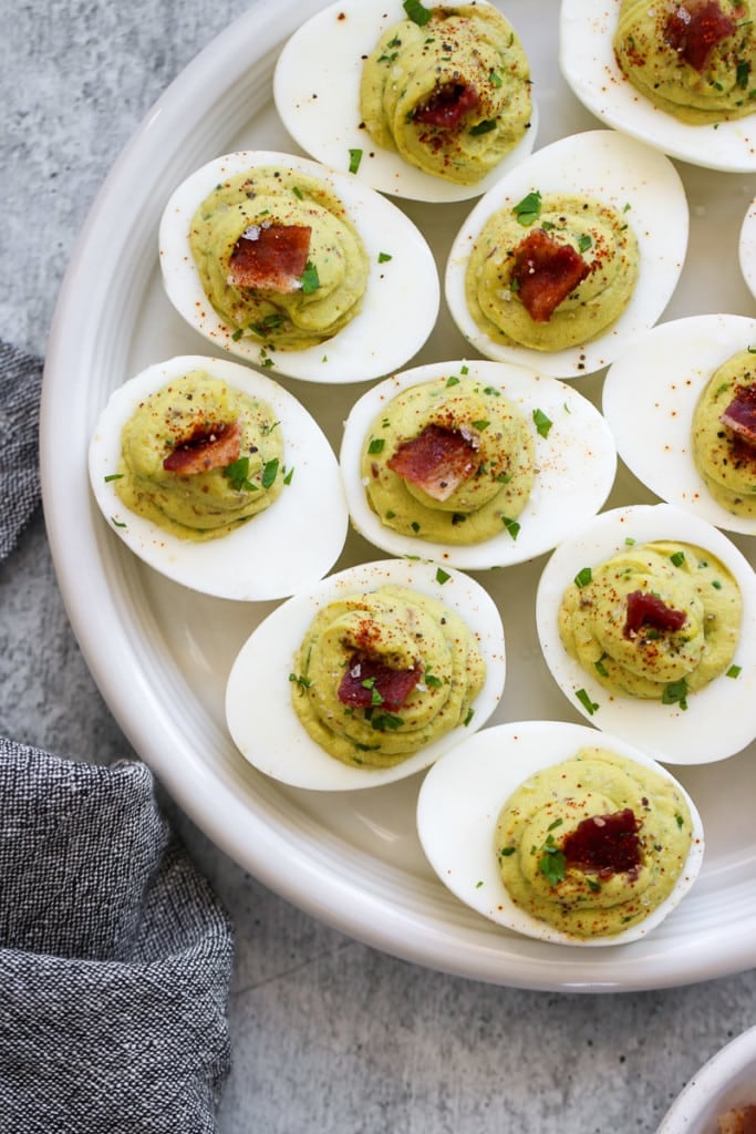 Overhead view of avocado deviled eggs topped with herbs, paprika, and bacon on a serving platter.