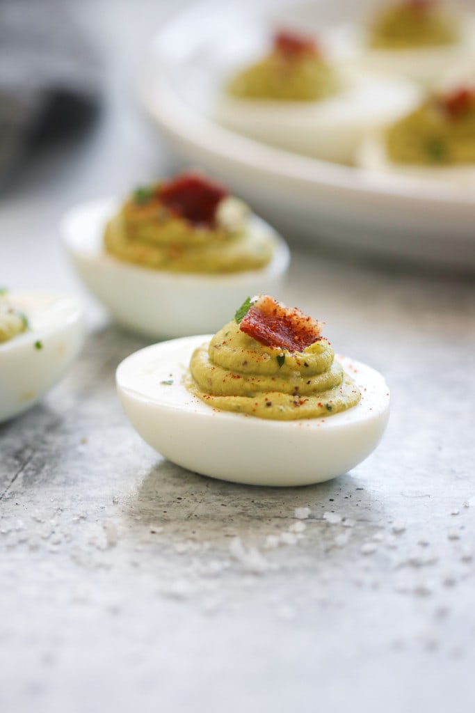 A single avocado deviled egg filled with creamy green avocado filling and topped with a little pieces of bacon.