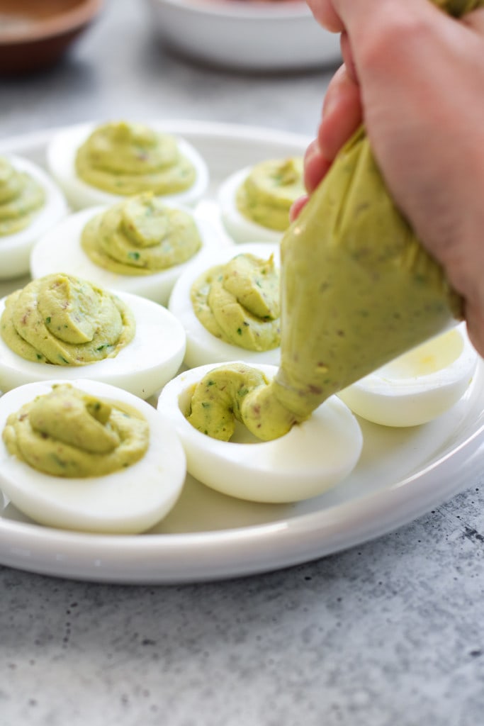 Avocado Deviled Eggs being filled with a piping bag filled with avocado filling piping them into the egg white shells. 