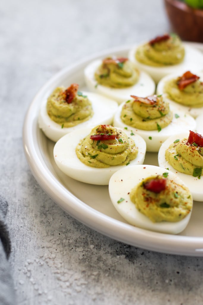 Avocado deviled eggs filled with creamy avocado filling and topped with a small piece of bacon, on a white serving tray.