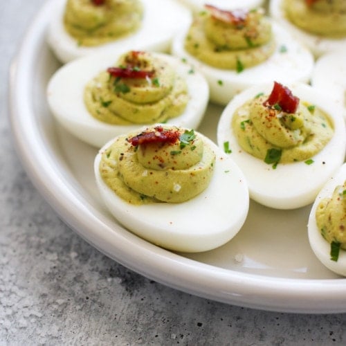 A white platter filled with avocado deviled eggs topped with little pieces of bacon.