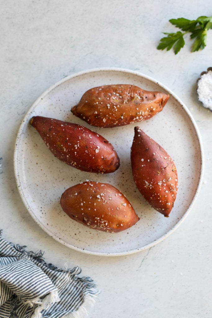 Four oven baked sweet potatoes coated in coarse sea salt on a speckled plate.