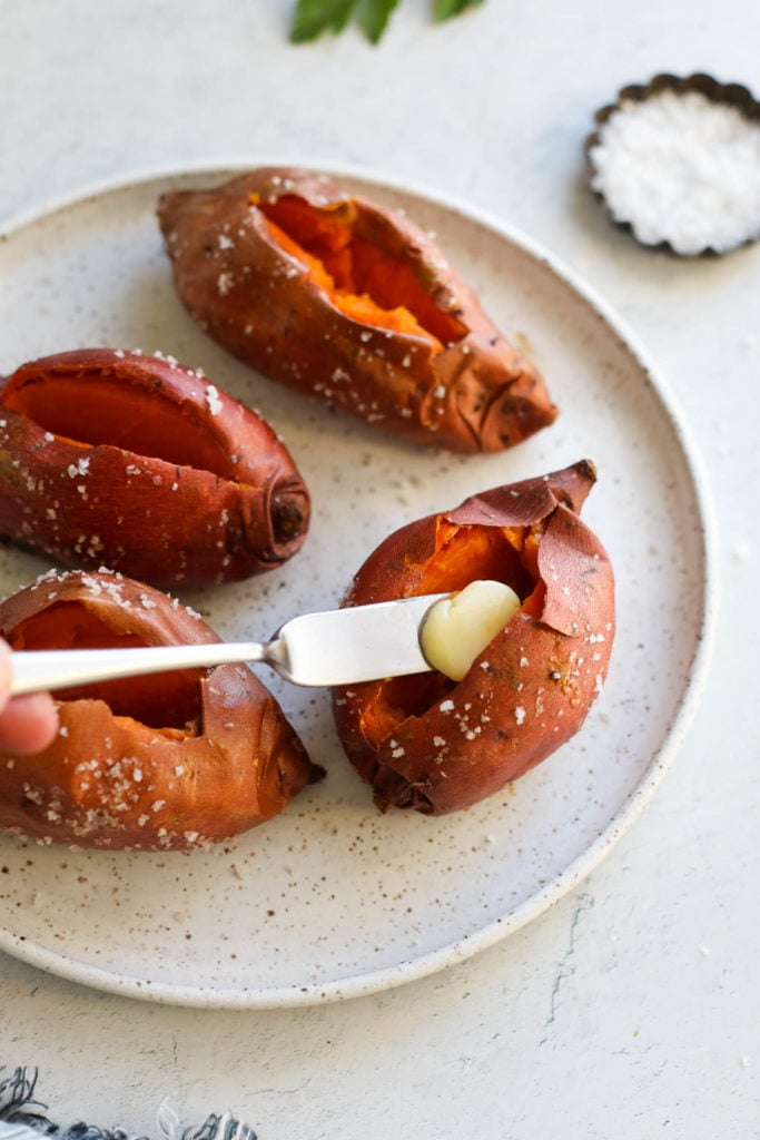 Four baked sweet potatoes on a speckled plate, cut lengthwise, with a pat of butter on a knife sliding into one cut sweet potato.