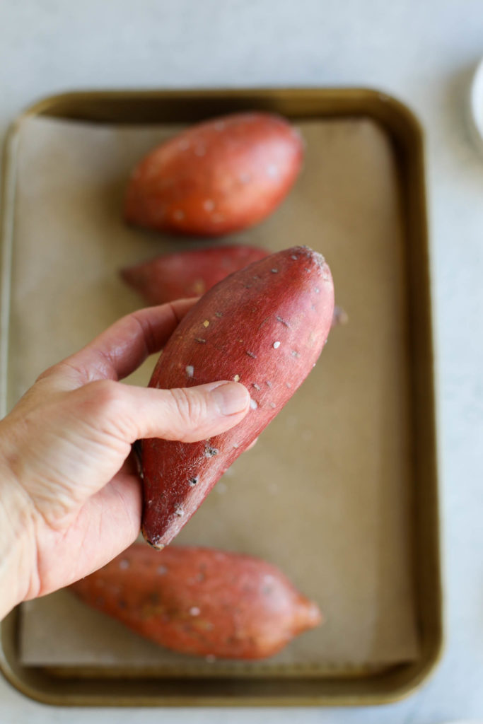 A hand holding a sweet potato over a parchment-covered baking sheet prepping for baking
