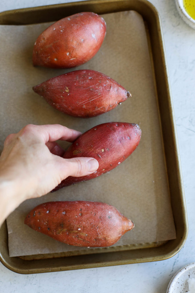 A hand placing an oiled sweet potato on a parchment-covered baking sheet holding three other sweet potatoes.