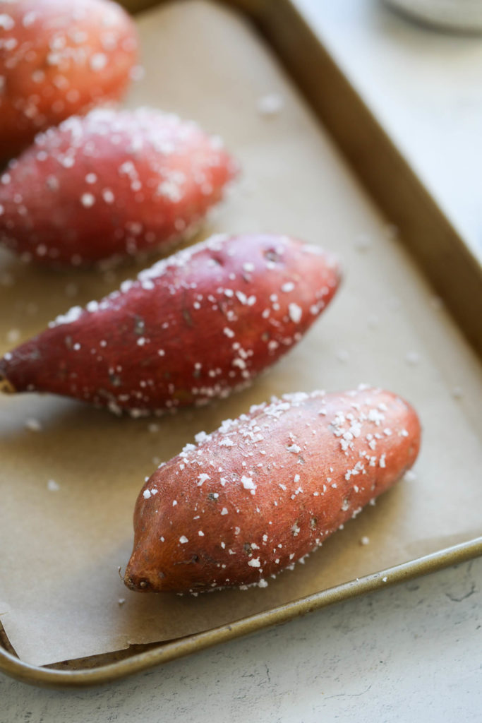 Four oiled sweet potatoes coated with coarse sea salt on a parchment covered baking sheet ready for the oven.