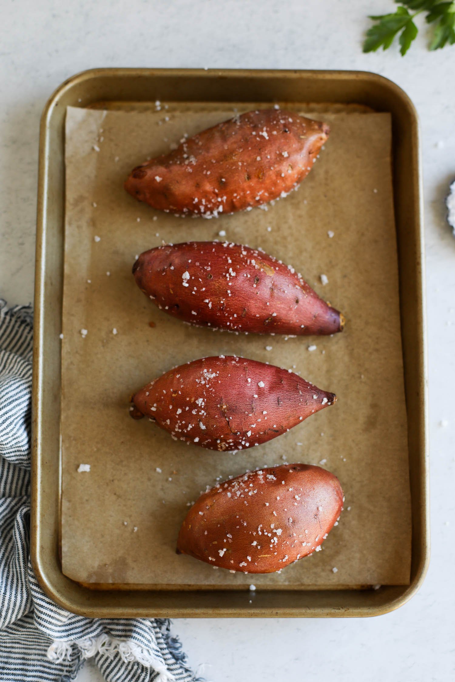 Overhead view of four baked sweet potatoes on a parchment covered baking sheet sprinkled with coarse sea salt.