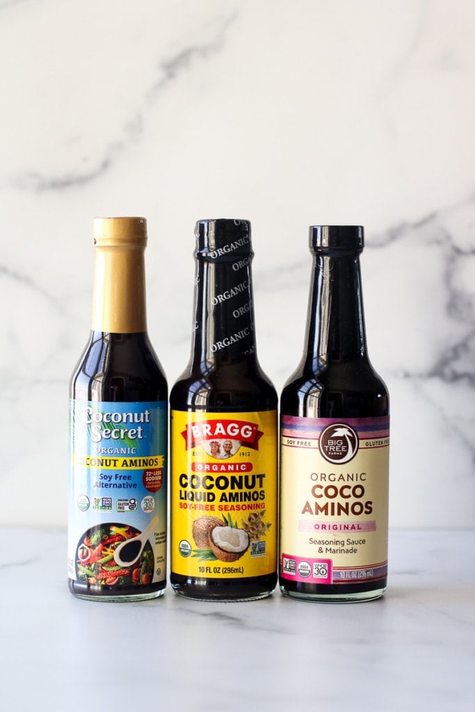 Three glass bottles of coconut aminos, in Coconut Secret brand, Bragg brand, and Big Tree brand labels. 