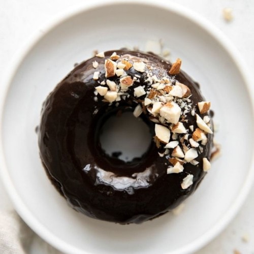 Overhead view of one flourless chocolate donut topped with chocolate icing and crushed almonds on a white plate.