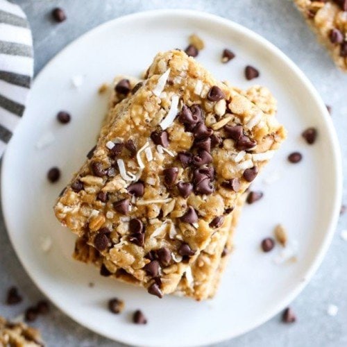 Three homemade chocolate chip clif bars stacked up on each other on a white plate, sprinkled with chocolate chips and coconut.