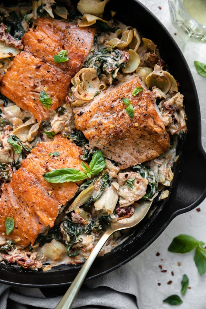 Overhead view of skillet creamy Tuscan salmon with artichoke hearts, spinach, and sun-dried tomatoes in a cast iron skillet with a gold spoon in the creamy vegetables. 