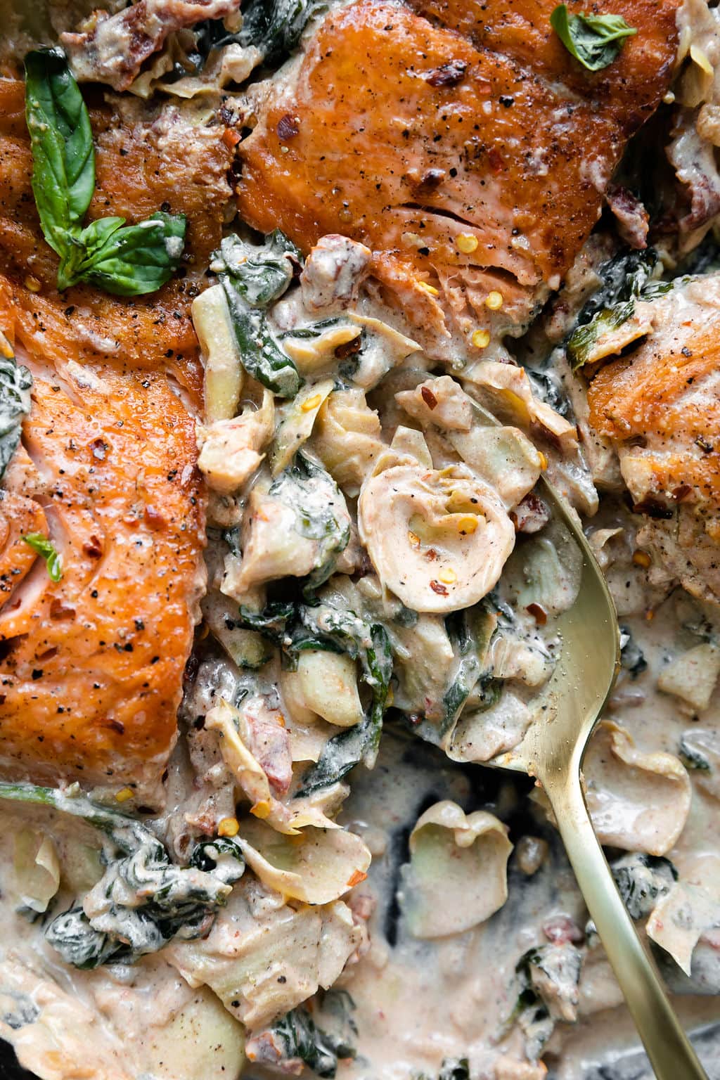 Close up view of creamy Tuscan skillet with artichoke hearts, spinach, and sun-dried tomatoes, a gold spoon scooping out a serving.