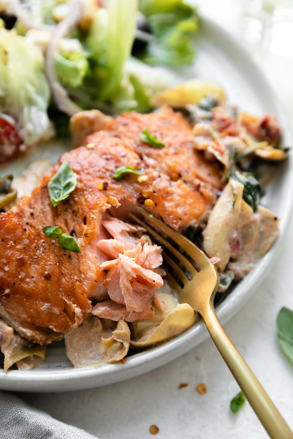 Close up view of a salmon fillet flaked with a gold fork, plated on a ceramic plate with creamy tuscan skillet of artichoke hearts, spinach, basil, and sun-dried tomatoes.