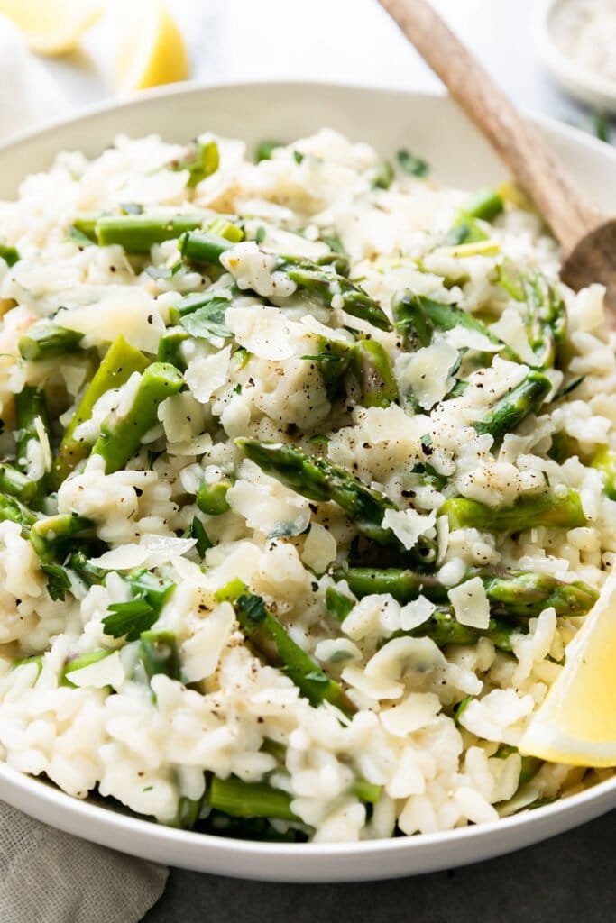 Close up view of lemon asparagus risotto in a white bowl with a wooden spoon, black cracked pepper and parmesan shavings sprinkled on top. 