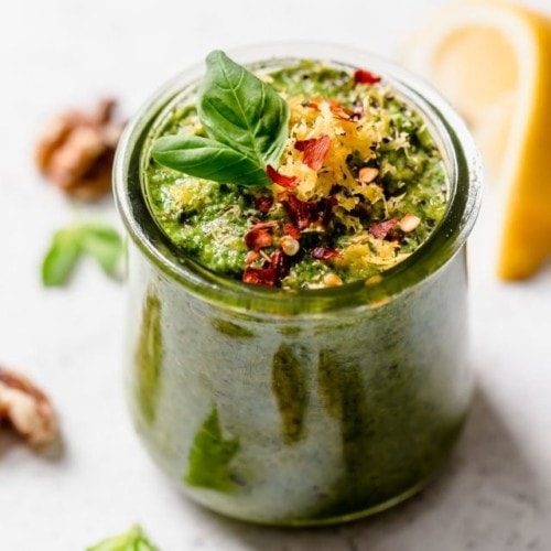 Basil Walnut Pesto in a short glass jar topped with small basil leaf, red pepper flakes, and lemon zest.