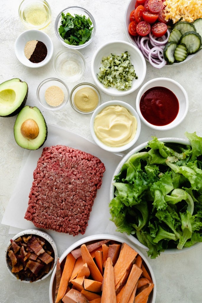 All ingredients for loaded burger bowls arranged in bowls on a counter. 