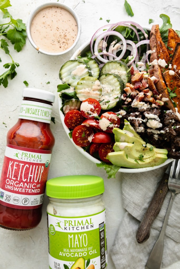 A loaded burger bowl filled with greens, hamburger, pickles, tomatoes, and sweet potato fries in a white bowl drizzled with special sauce. A bottle of Primal Kitchen ketchup and mayo on the side.